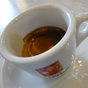 A white ceramic cup with a small amount of dark brown espresso sits on a white saucer, with a colorful logo on the cup. A glass spoon rests on the saucer beside the cup.