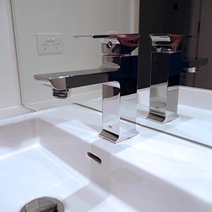 A modern chrome bathroom faucet is mounted on a white sink, with its reflection visible in the mirror behind it. An electrical outlet is seen on the wall above the sink.
