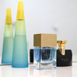 Three perfume bottles are displayed on a white surface, including a blue Gucci bottle with a gold cap, a black Bulgari bottle with a gold accent, and a tall blue-green Issey Miyake bottle with a yellow cap, reflected in a mirror.