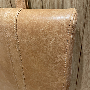Close-up of a tan-colored, leather-textured chair back with visible stitching, set against a light wood-paneled background.