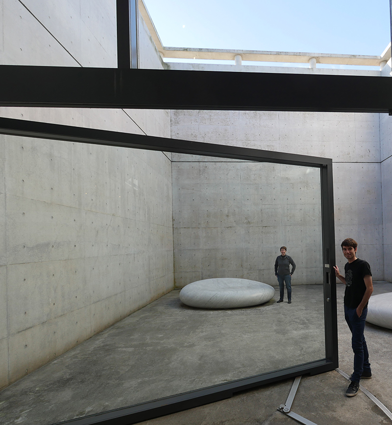 A man stands at an open glass door leading to a minimalist concrete courtyard with a circular stone bench. Another person stands inside the courtyard. The setting is modern and filled with natural light.