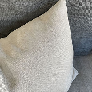 A close-up of a light beige pillow resting on a gray fabric sofa, showing the texture of both the pillow and the sofa upholstery.