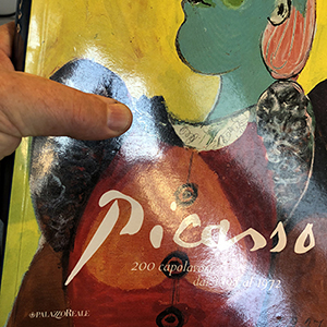 A hand holding a book with a colorful Picasso painting on the cover. The text reads “Picasso: 200 capolavori dal 1895 al 1972.” Bright yellow and abstract shapes are visible in the artwork.