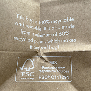 A brown paper bag with white text stating it is 100% recyclable and reusable, made from at least 60% recycled paper. The FSC logo and certification details are also visible.