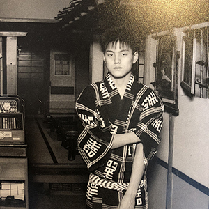 A young person in a patterned kimono stands in a hallway with traditional Japanese decor, looking at the camera with a neutral expression.