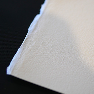 A close-up of the edge of a textured, off-white sheet of paper with a rough, torn side, set against a dark background.
