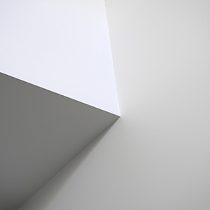 A minimalist image showing the corner of a white wall with sharp angles and soft shadows, creating geometric shapes against a light background.