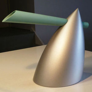 A modern, abstract metallic sculpture with a smooth, silver, cone-shaped base and a long, cylindrical, green extension angled through the top. The piece rests on a white surface against a dark background.