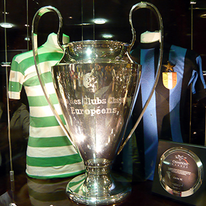 A large silver trophy with two handles is displayed between two classic soccer jerseys, one green and white striped and one blue and black striped, in a glass case.