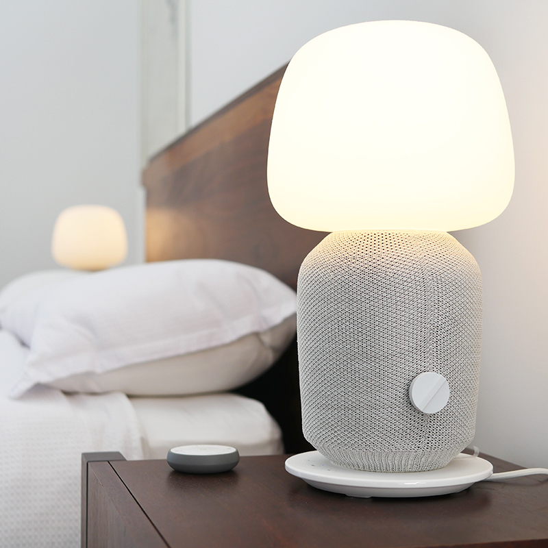 A modern bedside table lamp with a knitted grey base and glowing white shade sits on a wooden nightstand next to a bed with white linens; another similar lamp is visible in the background.