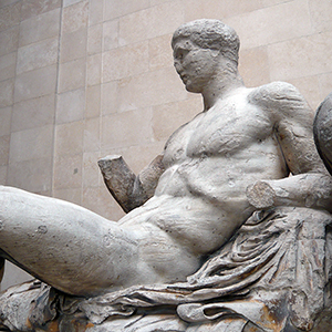 A weathered ancient marble statue of a reclining, muscular male figure with missing arms; the sculpture rests against draped fabric, positioned in front of a stone wall background.