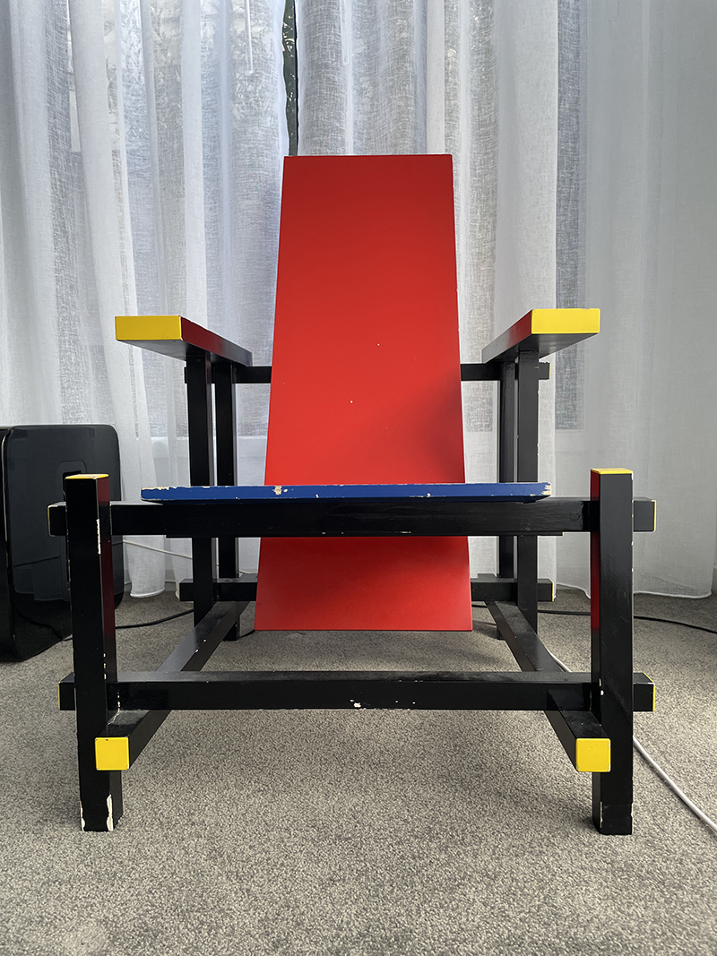 A geometric armchair with a red backrest, blue seat, and black frame accented with yellow edges, positioned on a carpeted floor in front of sheer white curtains.