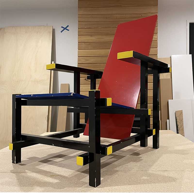 A modern chair with a black frame, yellow accents, a red angled backrest, and a blue seat, is set in a workshop with wooden panels and unfinished walls in the background.