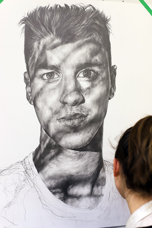 A person with brown hair observes a large, realistic pencil portrait of a man’s face, partially covered in shadow patterns, while working on its unfinished lower section.