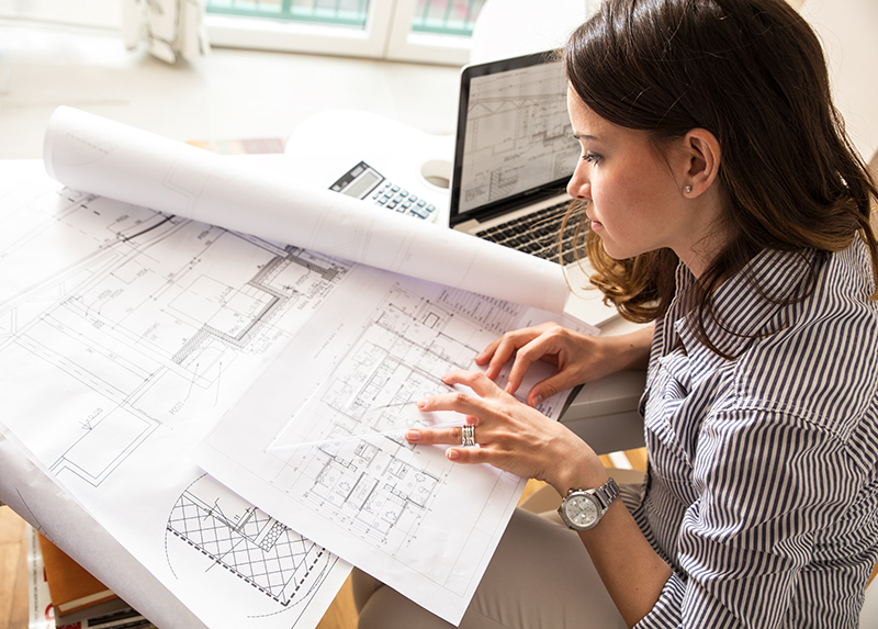 A woman reviews architectural blueprints at a desk with a laptop, calculator, and plans spread out in front of her, appearing focused on her work.