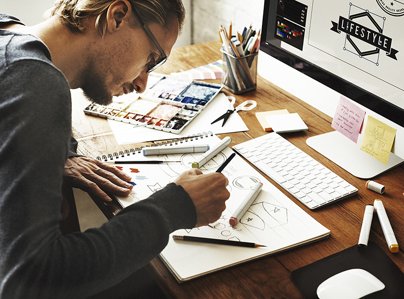 A man with glasses sketches geometric designs in a notebook at a wooden desk, surrounded by markers, a keyboard, and a computer displaying a logo that reads LIFESTYLE.