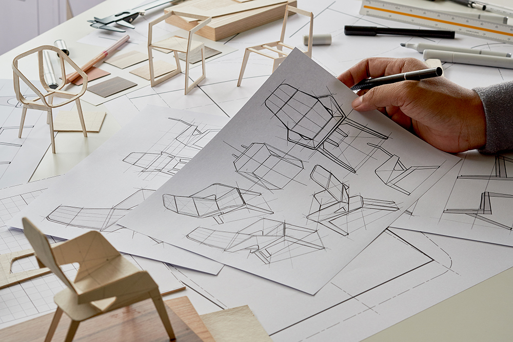 A hand draws chair design sketches on paper at a cluttered desk covered with other sketches, scale models of chairs, a ruler, and drawing tools.