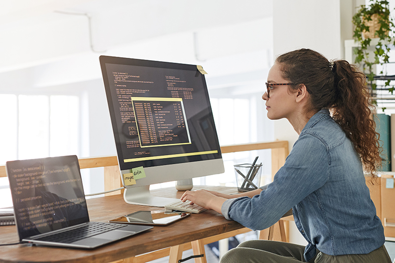 A woman with curly hair and glasses sits at a desk, working on a desktop computer displaying code. A laptop with code on the screen is next to her. The workspace is bright and modern with plants.