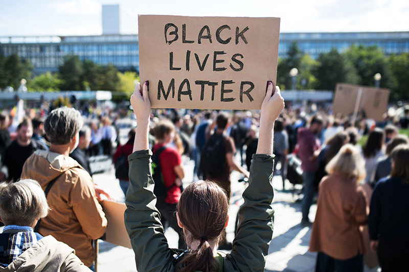 A person holds up a cardboard sign reading Black Lives Matter at a crowded outdoor protest, with many people gathered and a large building visible in the background.