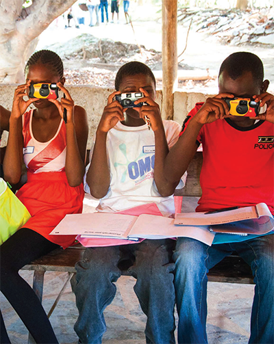 Three children sit on a bench holding disposable cameras up to their faces, aiming at the photographer. Notebooks and papers rest on their laps. They are outdoors under a wooden shelter.