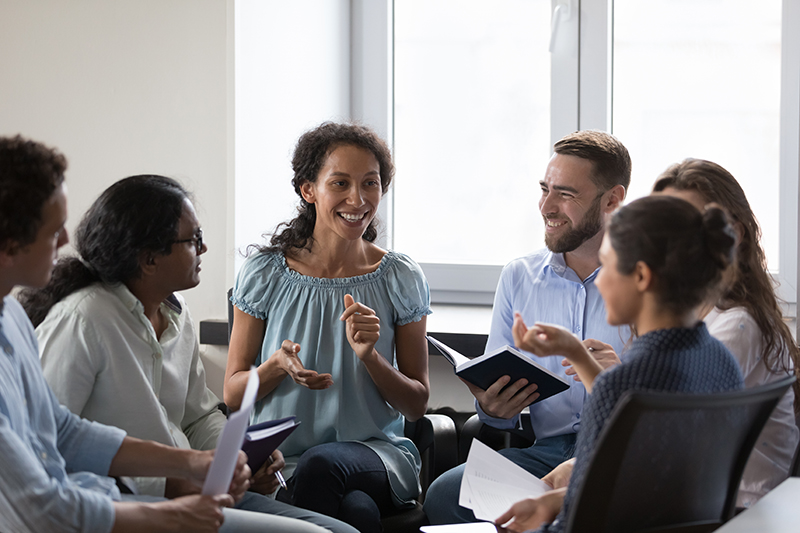 A diverse group of five adults sits in a circle indoors, engaged in a lively discussion. One woman in the center is smiling and speaking while others listen, smile, and hold notebooks or papers.