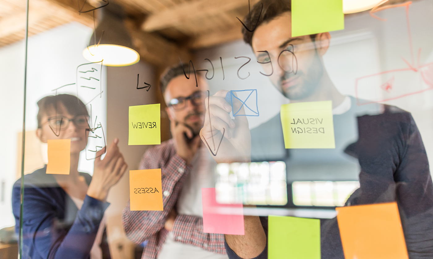 Three people brainstorm and write notes on a glass wall covered with colorful sticky notes and sketches, collaborating in a bright, modern office setting.