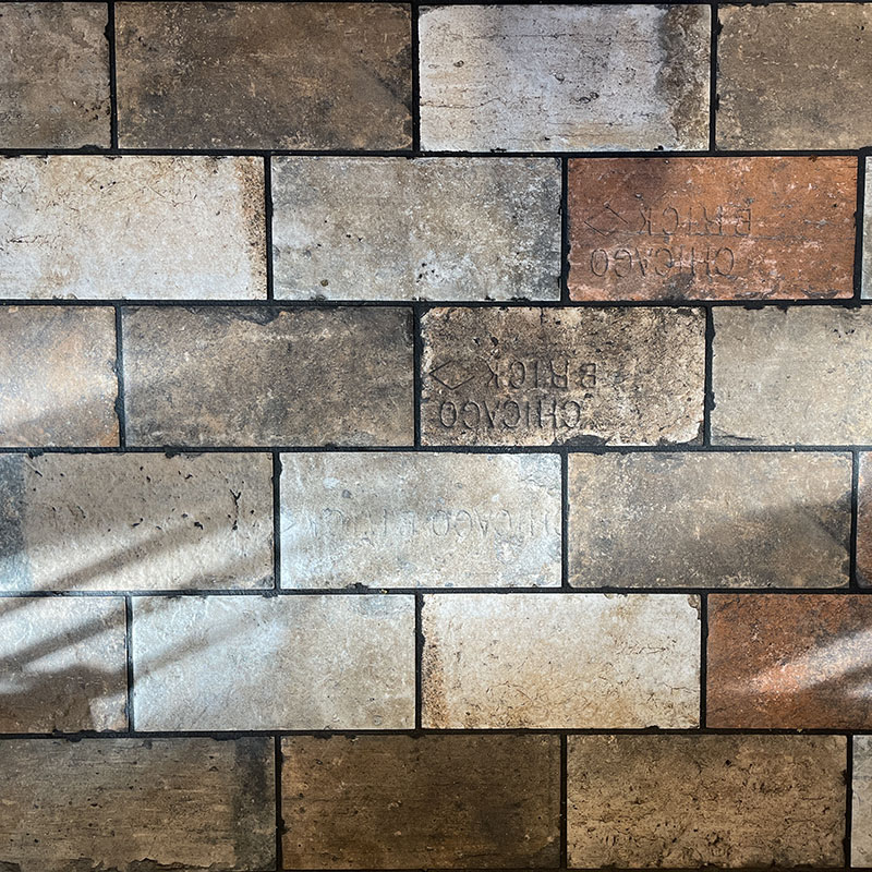 Rectangular tiles in earth tones form a brick pattern on a floor; some tiles have faint markings or text. Sunlight casts diagonal shadows across the tiles.