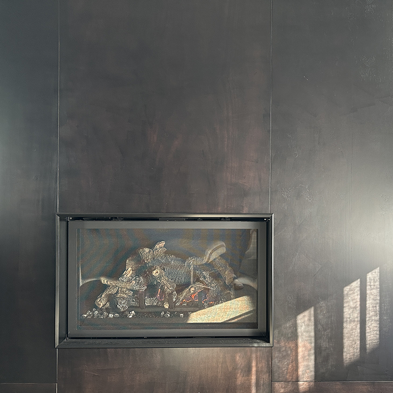 A modern, dark-colored fireplace with logs and decorative stones inside, set into a smooth, dark wall. Sunlight casts rectangular shadows on the wall to the right.