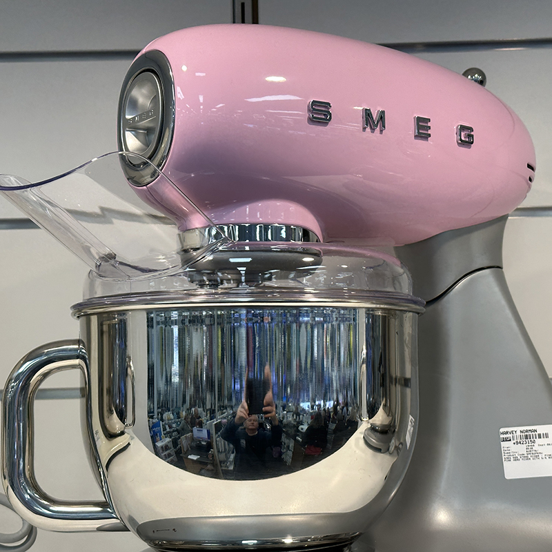 A pink SMEG stand mixer with a shiny metal mixing bowl sits on display. The bowl’s surface reflects the photographer and store interior. A white price sticker is visible on the base.