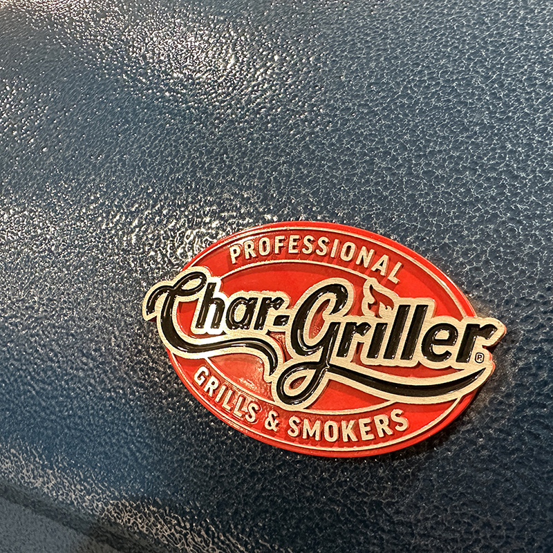 Close-up of a Char-Griller Professional Grills & Smokers logo on a textured, dark-colored grill surface. The logo is red, white, and gold with flame and smoke design elements.