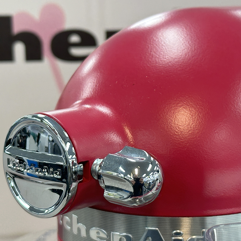 Close-up of a shiny, red KitchenAid stand mixer with chrome accents. Part of the KitchenAid logo and mixer head are visible against a blurred background with black letters.
