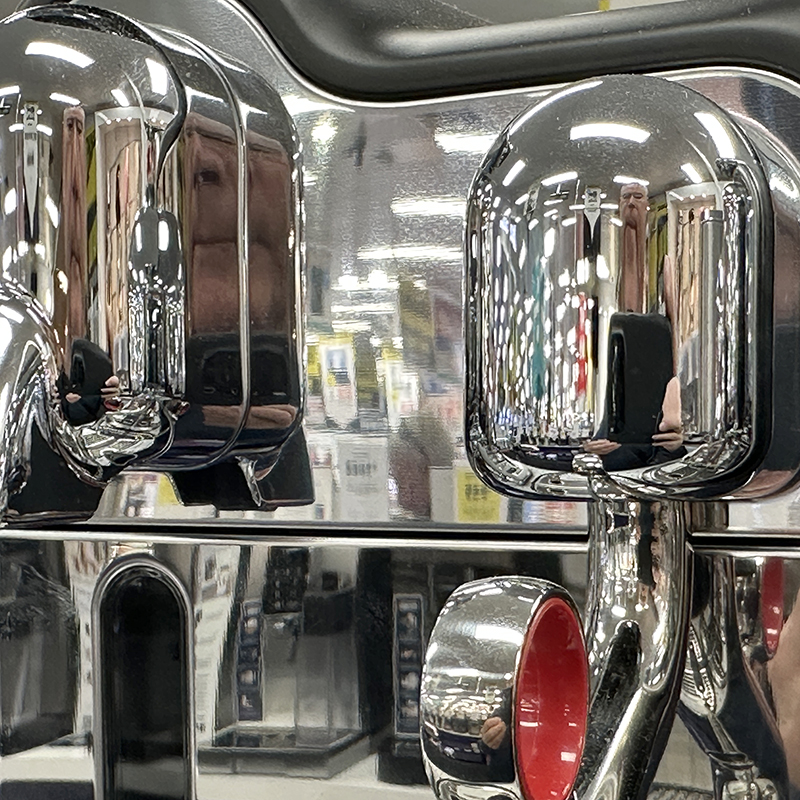 Close-up of a shiny chrome surface with multiple reflections of store shelves, ceiling lights, and people. Red and black details are visible among the metallic parts. The background is blurred with various signs and products.