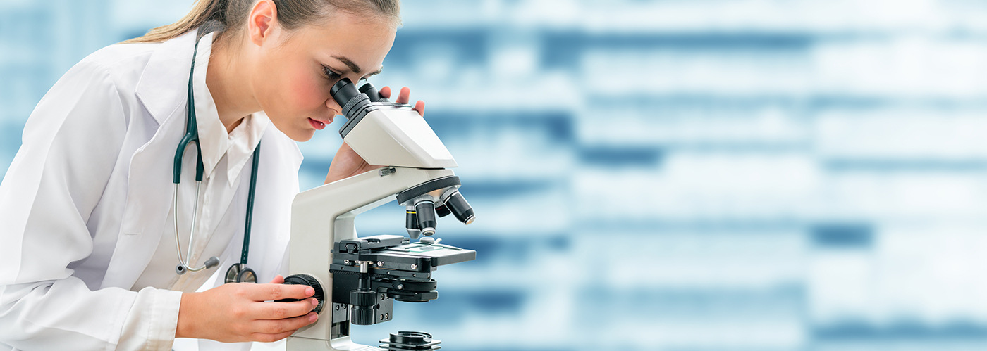 A scientist in a white lab coat and stethoscope looks into a microscope, focusing on a sample in a laboratory setting with blurred shelves in the background.