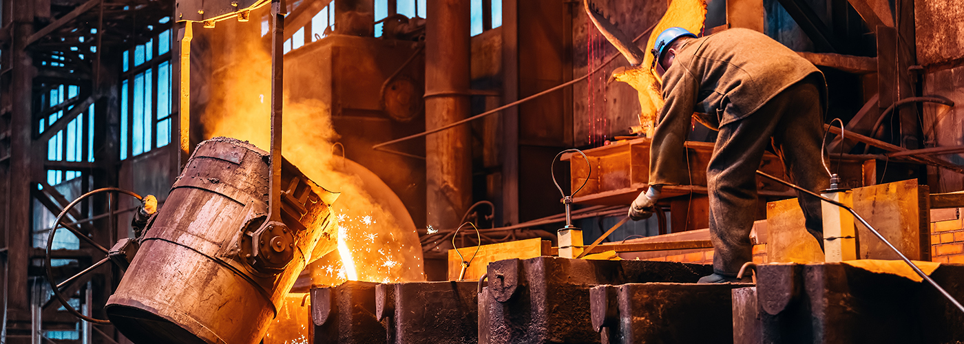 A worker in protective gear and a blue helmet pours molten metal from a large container in an industrial foundry, with sparks and glowing orange light illuminating the scene.