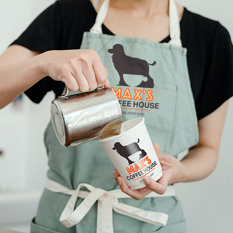 A barista wearing a green apron with a poodle logo and Maxs Coffee House text pours steamed milk into a paper coffee cup with the same logo and text.