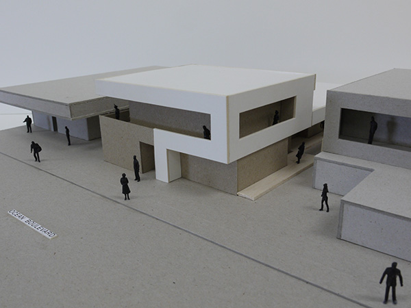 A minimalist architectural model with boxy, modern buildings in white and gray tones, surrounded by small black silhouette figures representing people.