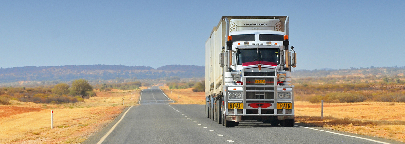 A large white semi-truck drives down a long, straight road through a dry, open landscape with low vegetation and distant hills under a clear blue sky.