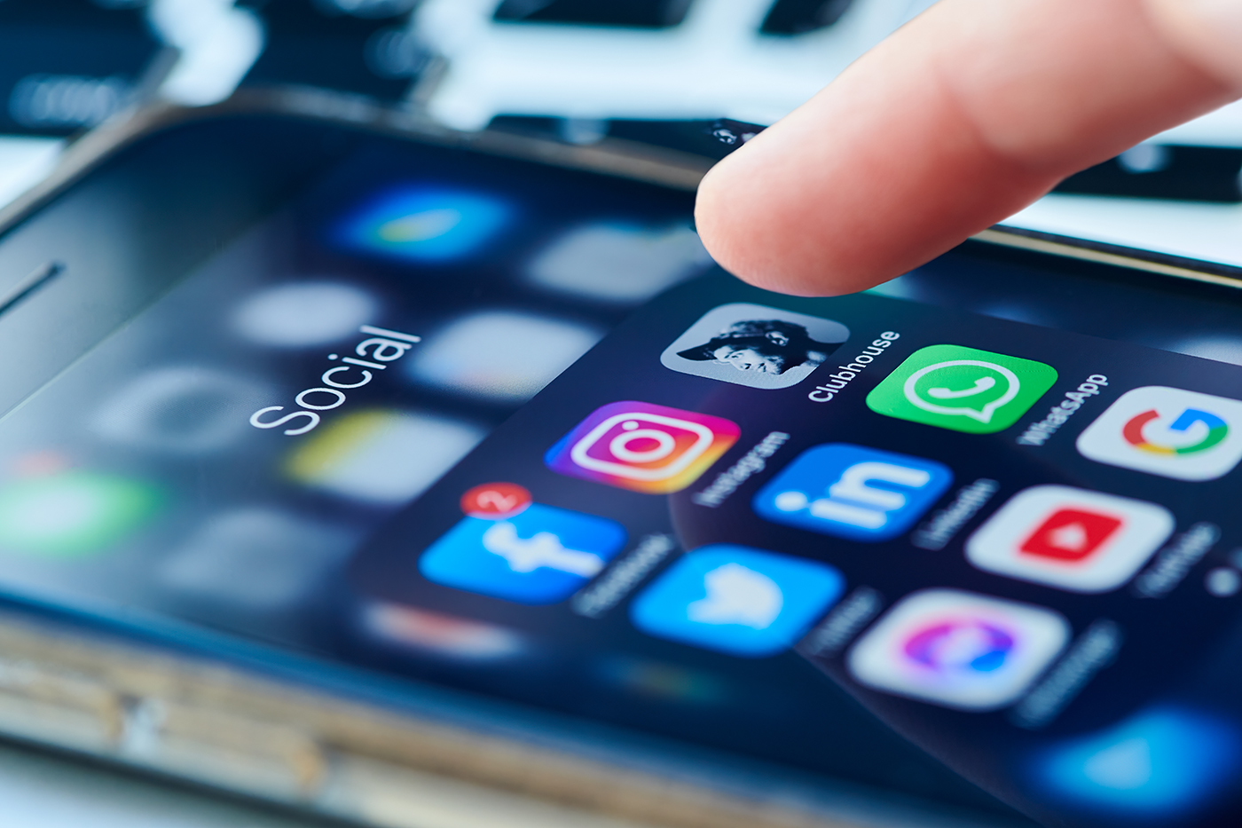 A close-up of a smartphone screen displaying social media apps, including Facebook, Twitter, Instagram, Clubhouse, WhatsApp, LinkedIn, and YouTube, with a finger about to tap the Clubhouse app icon.
