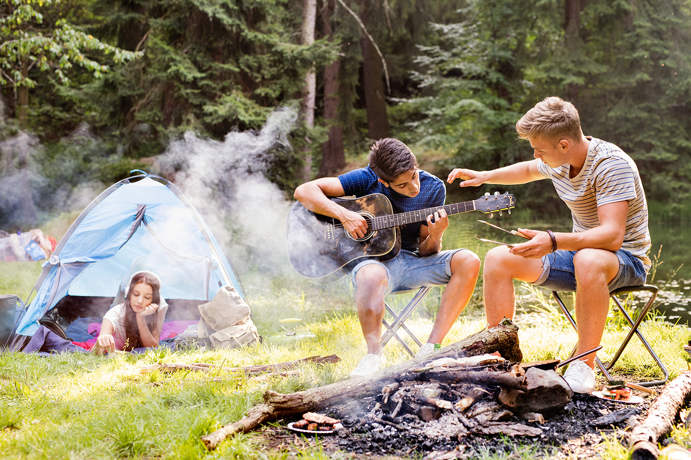 Two young men sit on camp chairs by a campfire in a forest; one plays guitar while the other gestures with roasting sticks. A girl relaxes inside a blue tent in the background. Smoke rises from the campfire.