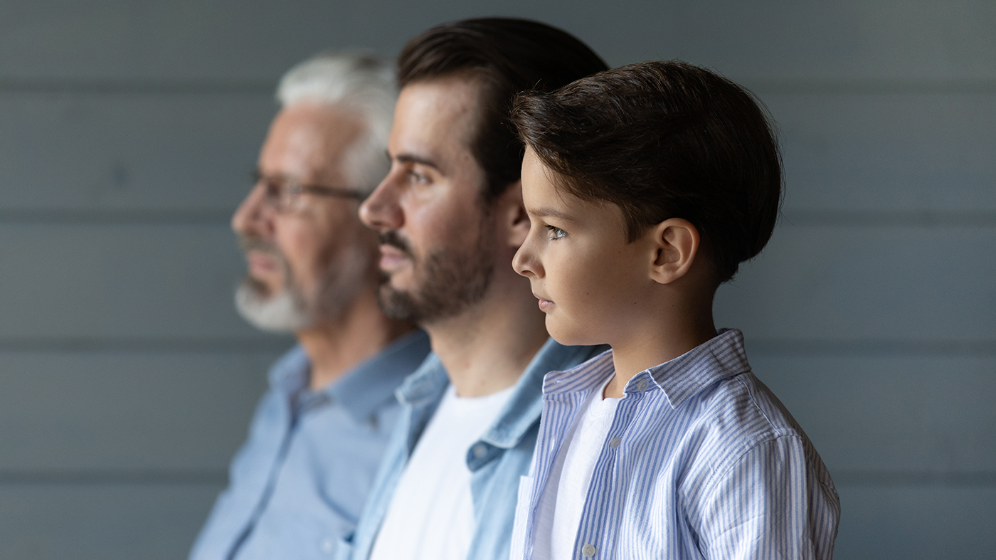 Three generations of men—a boy, a younger man, and an older man—stand in profile, facing right. All are wearing light blue shirts, and the background is a neutral-colored wall with horizontal panels.