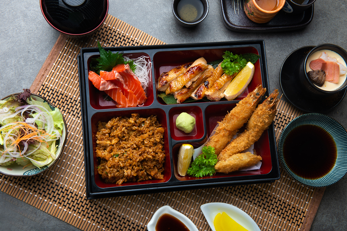 A Japanese bento box with fried shrimp, grilled chicken, salmon sashimi, rice, lemon wedges, wasabi, and garnish. Chawanmushi, salad, dipping sauces, and tea are arranged around the box on a woven mat.