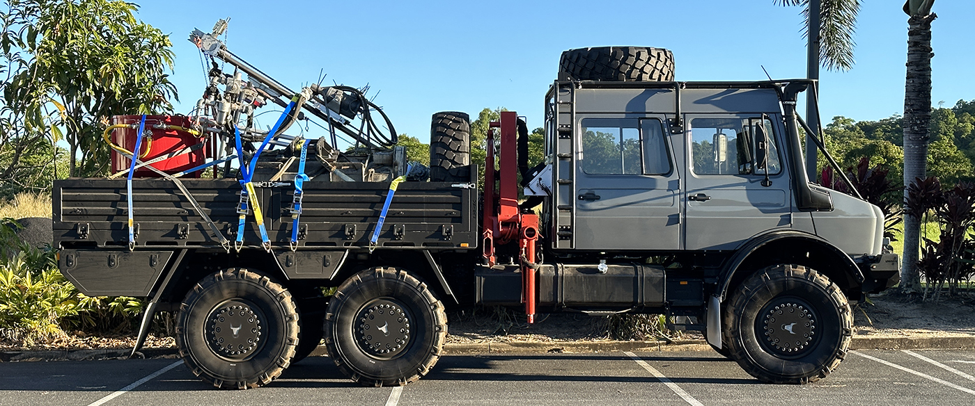 A large, gray six-wheeled truck is parked in a lot, loaded with equipment and spare tires in its open cargo bed, surrounded by tropical plants and trees on a sunny day.