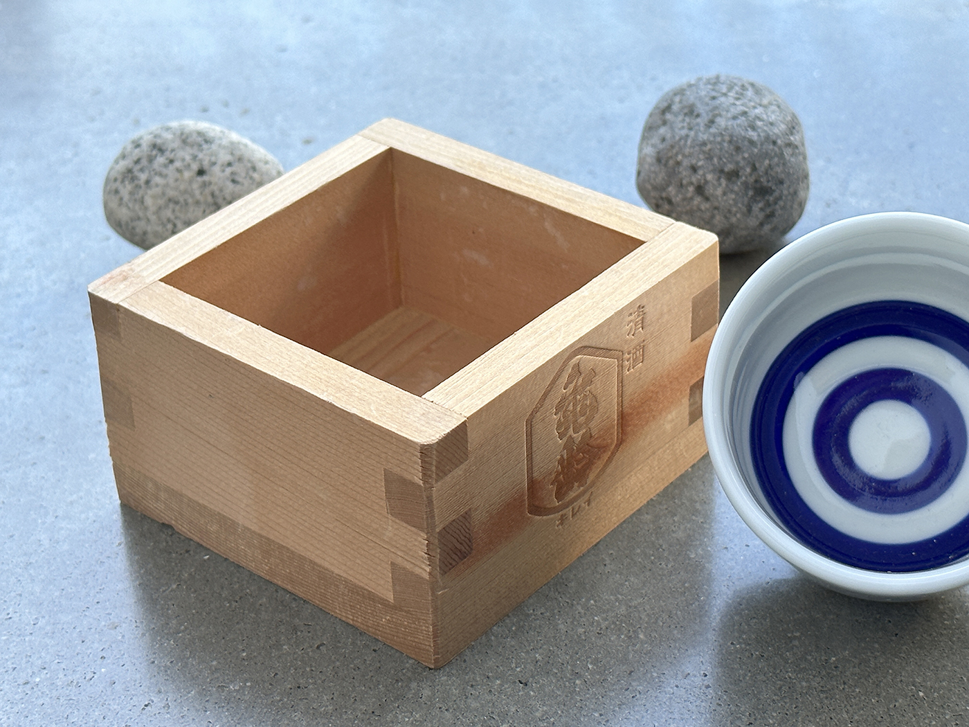 A wooden masu box sits on a gray surface beside a white ceramic sake cup with blue concentric circles inside. Two smooth gray stones are in the background.