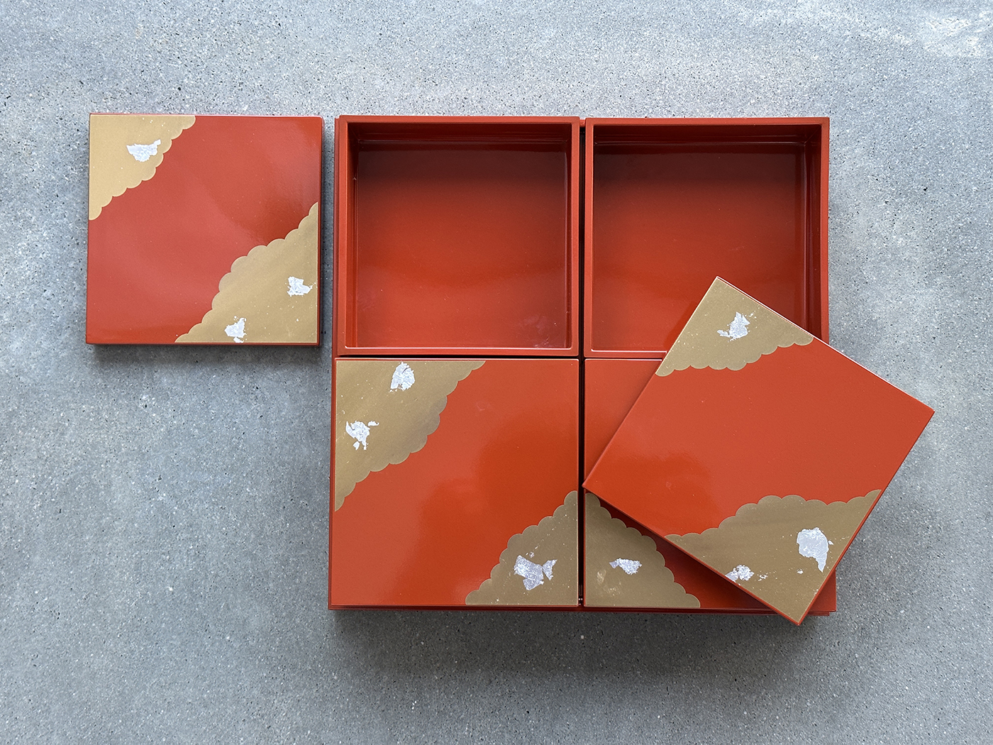 Four square lacquered boxes with red, gold, and white patterns are arranged on a gray surface. Two boxes are open, revealing empty interiors, and two lids are set aside, showing the decorative design.