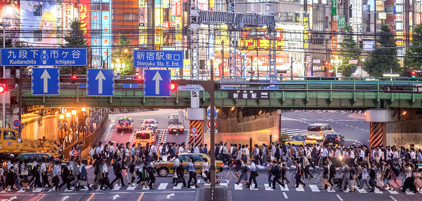 A busy city street in Tokyo with many people crossing the road at a crosswalk, cars driving underneath a green overpass, and colorful illuminated signs on buildings in the background.