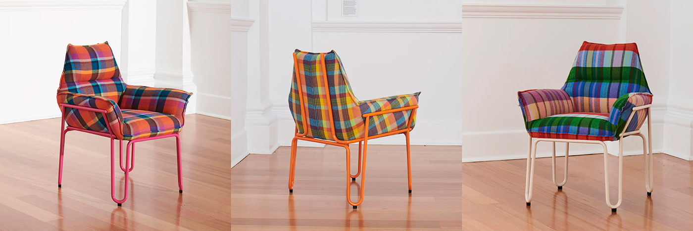 Three colorful armchairs with plaid-patterned cushions and metal frames in pink, orange, and white are displayed on a wooden floor against a white wall.