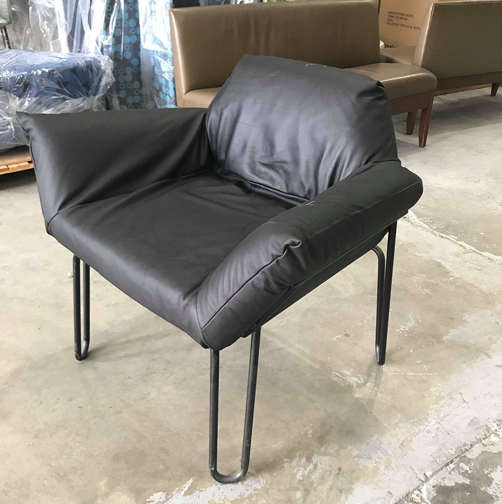 A black cushioned chair with thin metal legs sits on a concrete floor in a warehouse, surrounded by other furniture pieces and packaging materials.