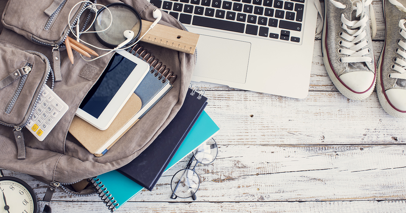 A backpack with school supplies, an open laptop, notebook, calculator, eyeglasses, ruler, earphones, tablet, and sneakers on a white wooden surface.