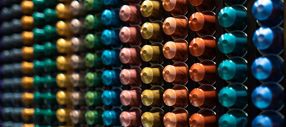 Rows of colorful coffee pods are neatly arranged in a grid pattern, with each row featuring pods of the same color, including green, yellow, brown, blue, and orange. The image is taken at an angle.