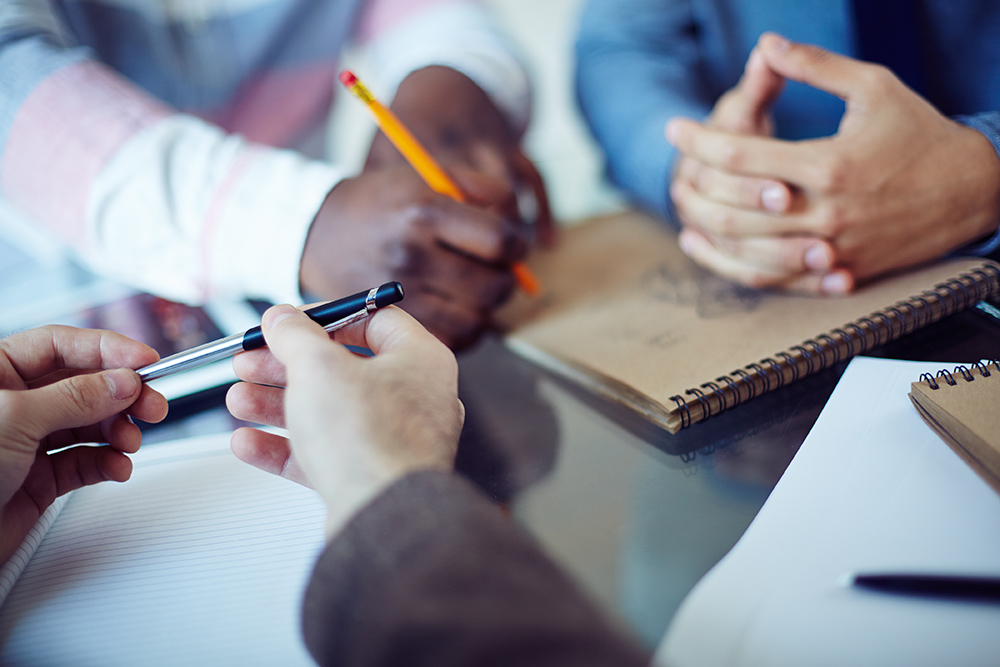 Three people sit around a table with notebooks, one writing with a pencil, another holding a pen, and the third resting clasped hands on a notepad, suggesting a collaborative discussion or meeting.